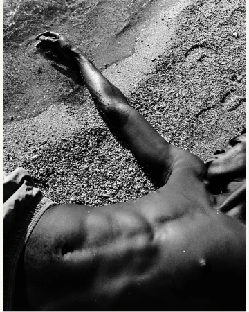 Monochrome photograph of an adult lying on a sandy beach with arm outstretched towards the ocean water lapping on the shore in daylight.