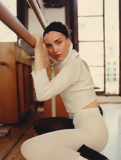 Indoor portrait of a woman in white turtleneck and leggings, kneeling on a wood floor with soft natural lighting from a nearby window.