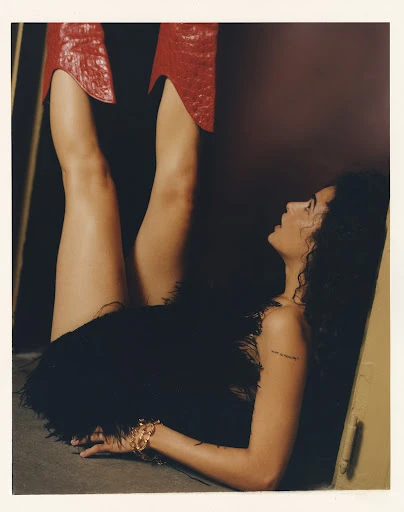 Woman with curly hair wearing a black, feathered skirt, laying on floor. Legs extend vertically with shiny, red, reptile-texture boots.
