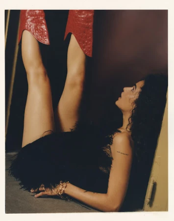 Woman with curly hair wearing a black, feathered skirt, laying on floor. Legs extend vertically with shiny, red, reptile-texture boots.