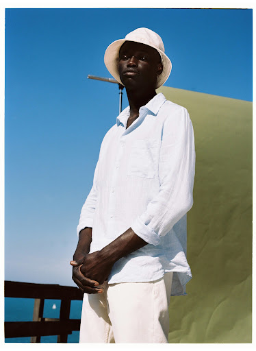 Studio shot of a Black male model in a linen shirt and bucket hat, posing outdoors against a blue sky and backdrop.