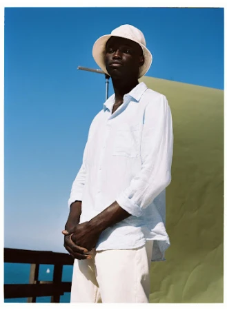 Studio shot of a Black male model in a linen shirt and bucket hat, posing outdoors against a blue sky and backdrop.