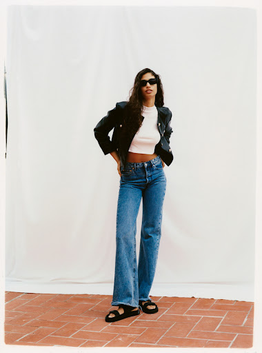 Model wearing dark sunglasses, black jacket, white crop top, and blue jeans standing against white backdrop.