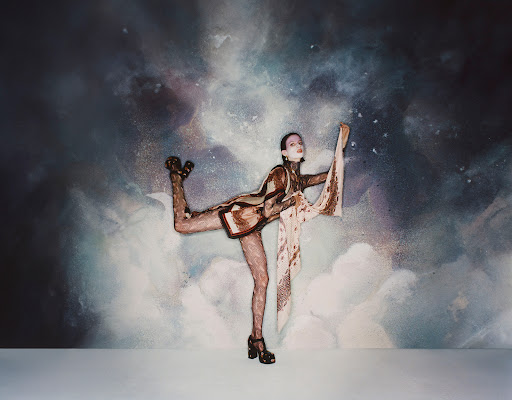 A slender female fashion model in a geometric-patterned outfit strikes an arabesque pose in front of an abstract celestial background.