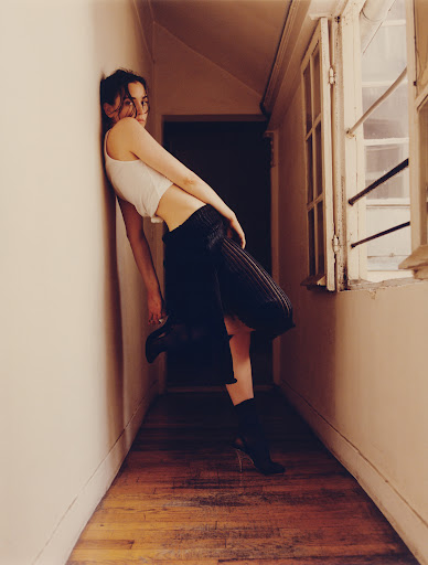 Young woman in white crop top and black skirt leaning against a wall in a hallway with a window, posing for a fashion-style photograph.