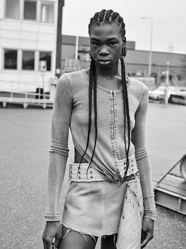 Black and white portrait of a young adult woman with braids, wearing a long sleeve top and skirt, posing outdoors on a paved surface.