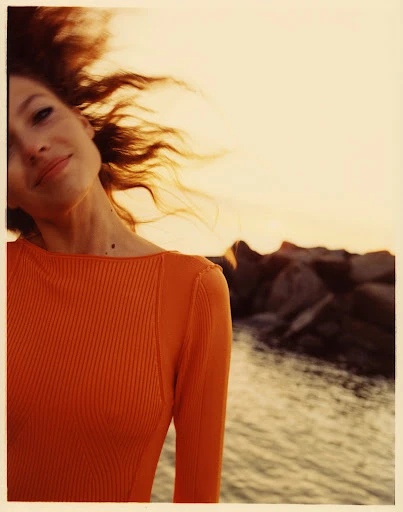 Portrait of a young adult woman with wavy brown hair and orange sweater, smiling softly near a rocky shore and tranquil water.