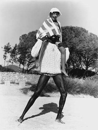 Black and white photo of an adult carrying a child on their shoulders on a sandy beach near trees and a rock wall.
