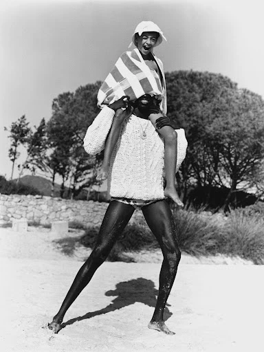 Black and white photo of an adult carrying a child on their shoulders on a sandy beach near trees and a rock wall.