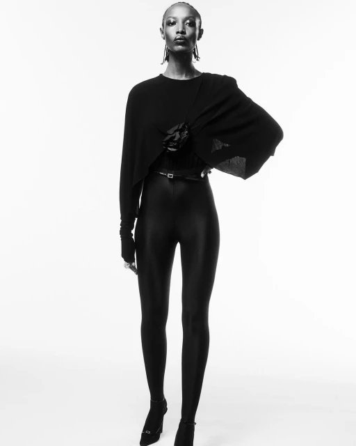 A full-length portrait in black and white showcases a female model in a draped top, leggings, heels, and statement earrings against a white backdrop.