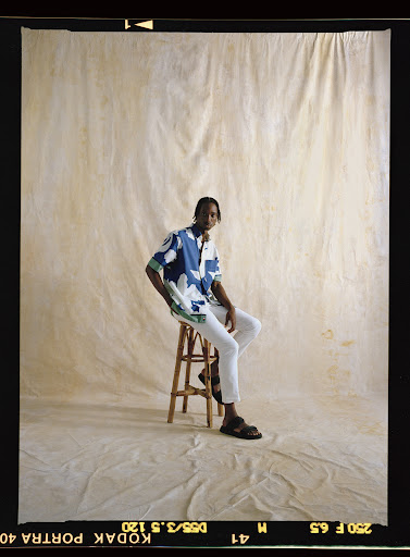 A dark-skinned man with a patterned shirt and white pants sits on a wooden stool in a studio with a neutral canvas backdrop.