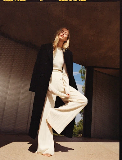 A fair-skinned woman with long blonde hair in cream-colored wide-leg pants and a sweater, topped with a black overcoat, poses in an urban setting.