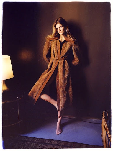 Woman in long brown coat poses indoors near lamp and bedframe, in muted lighting against a brown wall.