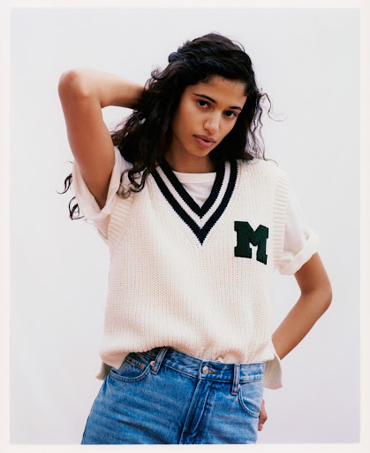 Woman wearing a knit varsity sweater vest and jeans stands in front of a solid white background in a medium studio shot.