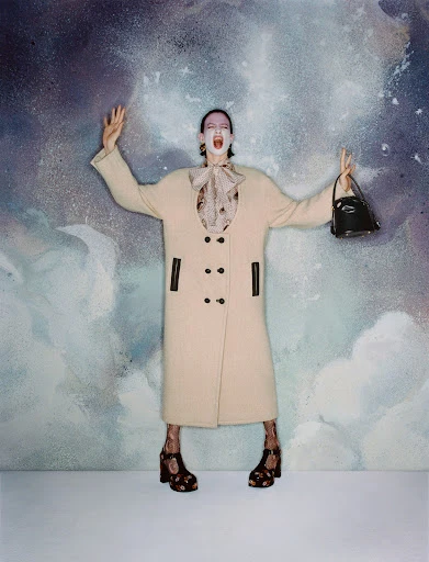 Fashion shot of a model posing in a cream coat, floral blouse, and sandals against an abstract cloud-like background, holding a purse.