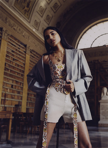 Adult female poses in a photo studio wearing a floral print top and light blue blazer, in front of bookshelves and architectural details.
