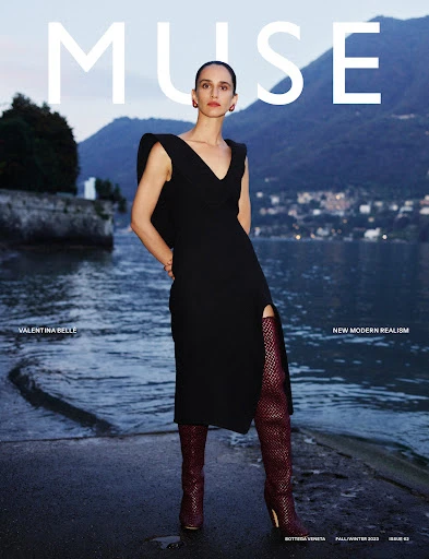 Magazine cover featuring a woman in a black dress and high boots standing by a lake with mountains in the background at dusk.