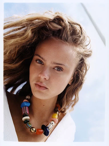 Portrait of a young adult female model with curly blonde hair wearing a colourful beaded necklace, against a bright, neutral background.
