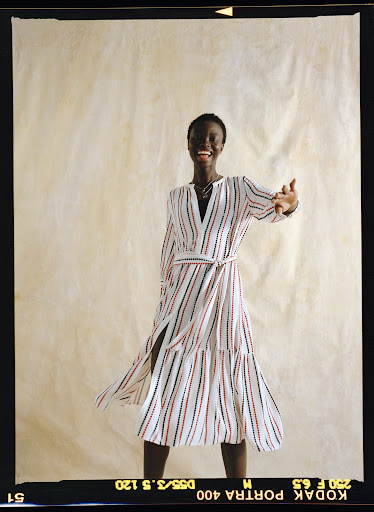 Smiling Black woman in striped wrap dress against cream paper backdrop. Fashion shot, waist-up, arms raised in greeting.