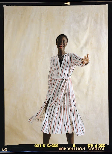 Smiling Black woman in striped wrap dress against cream paper backdrop. Fashion shot, waist-up, arms raised in greeting.