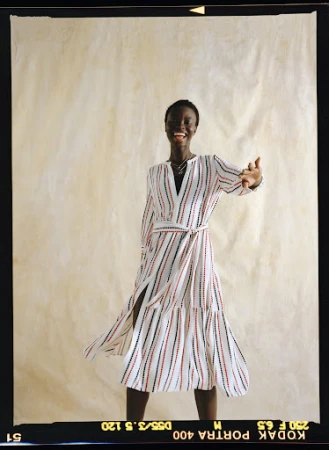 Smiling Black woman in striped wrap dress against cream paper backdrop. Fashion shot, waist-up, arms raised in greeting.