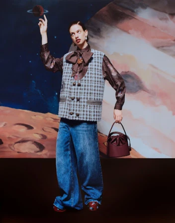 A woman in wide-legged jeans and a vest poses in front of a planetary backdrop, holding a maroon purse, with a moody studio setting.