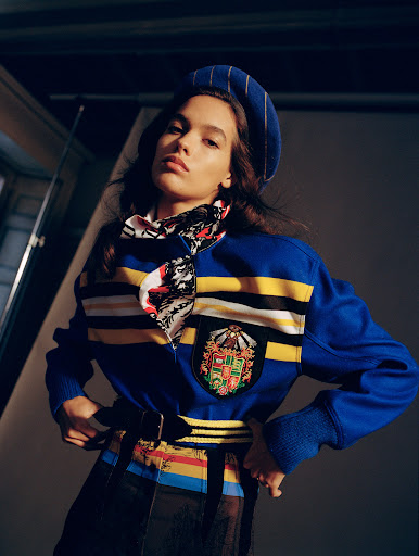 Brunette woman posing in a photo studio wearing a blue beret, patterned scarf, and blue striped sweater with a crest emblem.