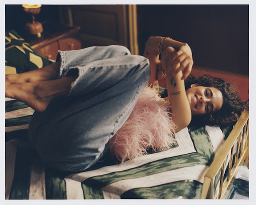 Young woman with curly hair lying on a bed wearing a pink feathered top and denim jeans photographed by Luca Campri