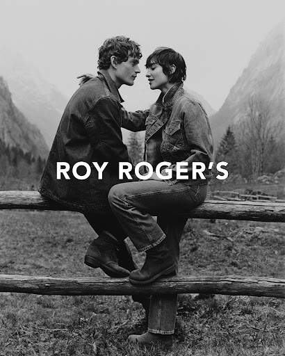 Black and white shot of a stylish young couple in denim jackets and boots in a mountain setting, next to a wooden fence.
