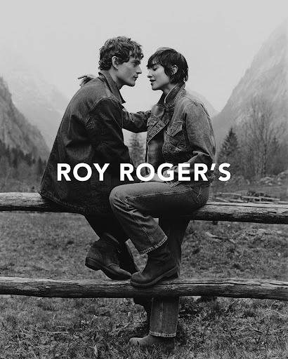 Black and white shot of a stylish young couple in denim jackets and boots in a mountain setting, next to a wooden fence.