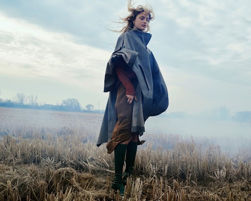 A blonde woman in a cape and knee-high boots stands in a harvested field under a cloudy sky, creating a dramatic, fashion-editorial look.