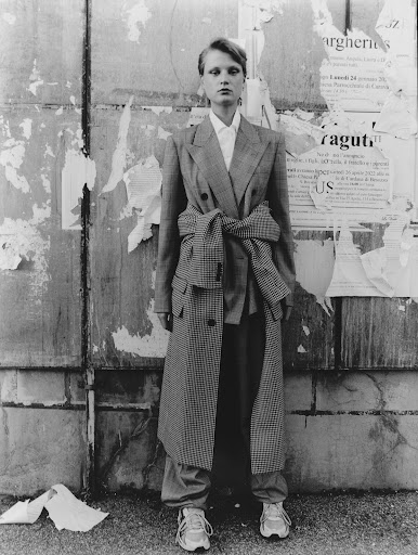 Black and white full shot of a female model in a plaid coat and pants standing in front of a weathered urban wall with ripped posters.