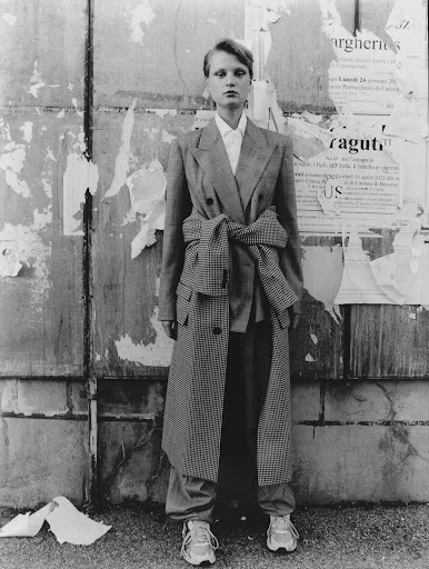 Black and white full shot of a female model in a plaid coat and pants standing in front of a weathered urban wall with ripped posters.
