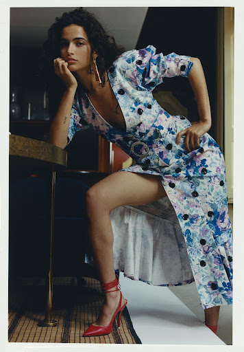 A brunette woman poses indoors wearing a floral print dress and red heels. She leans on a table with hand near her face.
