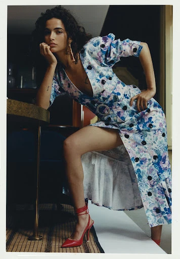 A brunette woman poses indoors wearing a floral print dress and red heels. She leans on a table with hand near her face.