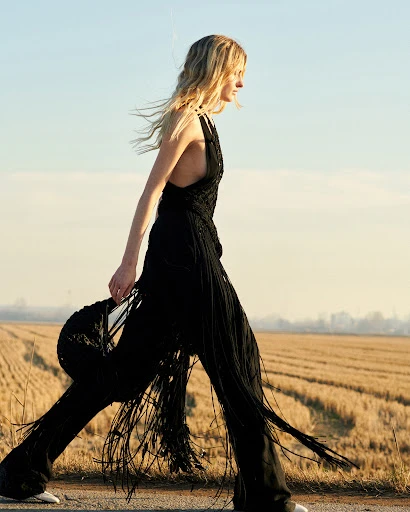 A woman with blonde hair walks along a rural road, wearing a long black fringed halter jumpsuit with a field in the background.