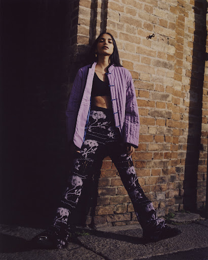 Woman poses in patterned pants and a jacket near a brick wall, featuring a blend of fashion and urban environment in natural light.