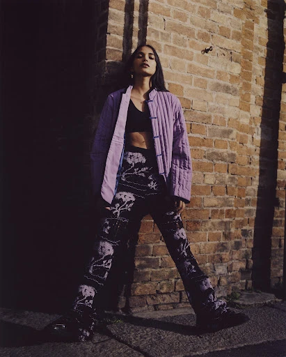 Woman poses in patterned pants and a jacket near a brick wall, featuring a blend of fashion and urban environment in natural light.