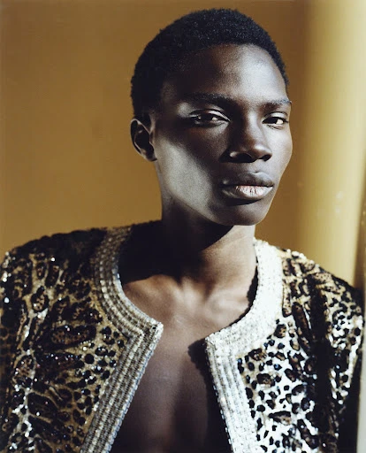 Close-up portrait of an adult male with dark skin wearing a leopard-print jacket against a yellow background in a photo studio.