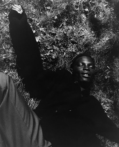 Black and white portrait of a young man lying on his back in grass with his arm raised, wearing a dark jacket in natural light.