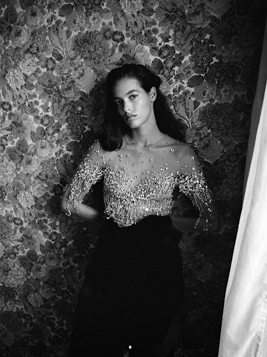 Monochrome portrait of a young woman with long dark hair in a beaded top posing against a floral wallpaper background with diffused light.