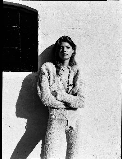 An adult female model with crossed arms poses in textured pants and sweater next to a window shadow against a concrete wall in black and white.