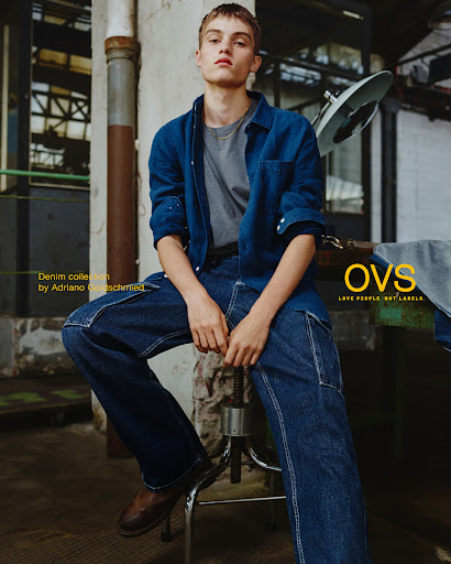 Man in blue denim shirt and jeans sits on a metal stool in a warehouse-style space, posing for a fashion editorial campaign.
