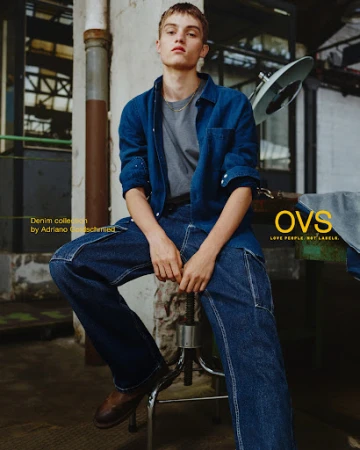 Man in blue denim shirt and jeans sits on a metal stool in a warehouse-style space, posing for a fashion editorial campaign.
