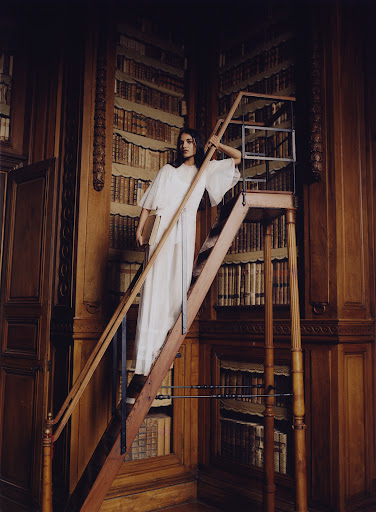 Woman in white dress on a library ladder, surrounded by dark wood bookshelves in a vintage library setting with natural light.