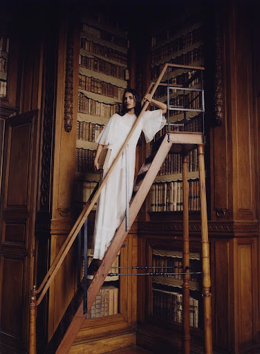 Woman in white dress on a library ladder, surrounded by dark wood bookshelves in a vintage library setting with natural light.