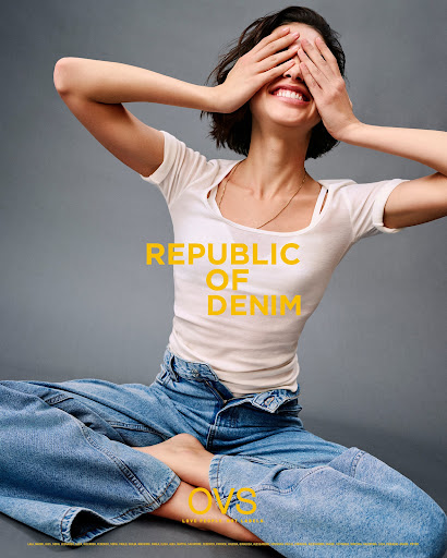 Smiling woman with short hair covers her eyes in fashion shot. She wears a white t-shirt and wide-leg jeans on a neutral background.