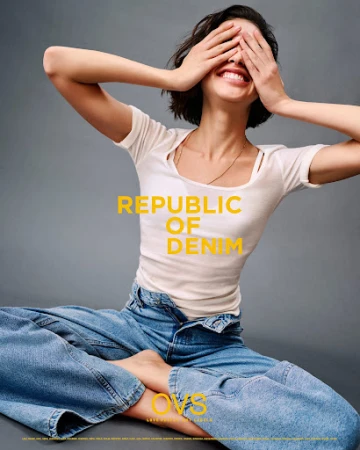 Smiling woman with short hair covers her eyes in fashion shot. She wears a white t-shirt and wide-leg jeans on a neutral background.
