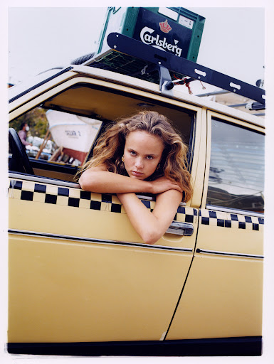 Portrait of a young woman with long hair leaning out the window of a vintage yellow car with black and white checkered trim.