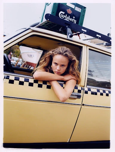 Portrait of a young woman with long hair leaning out the window of a vintage yellow car with black and white checkered trim.